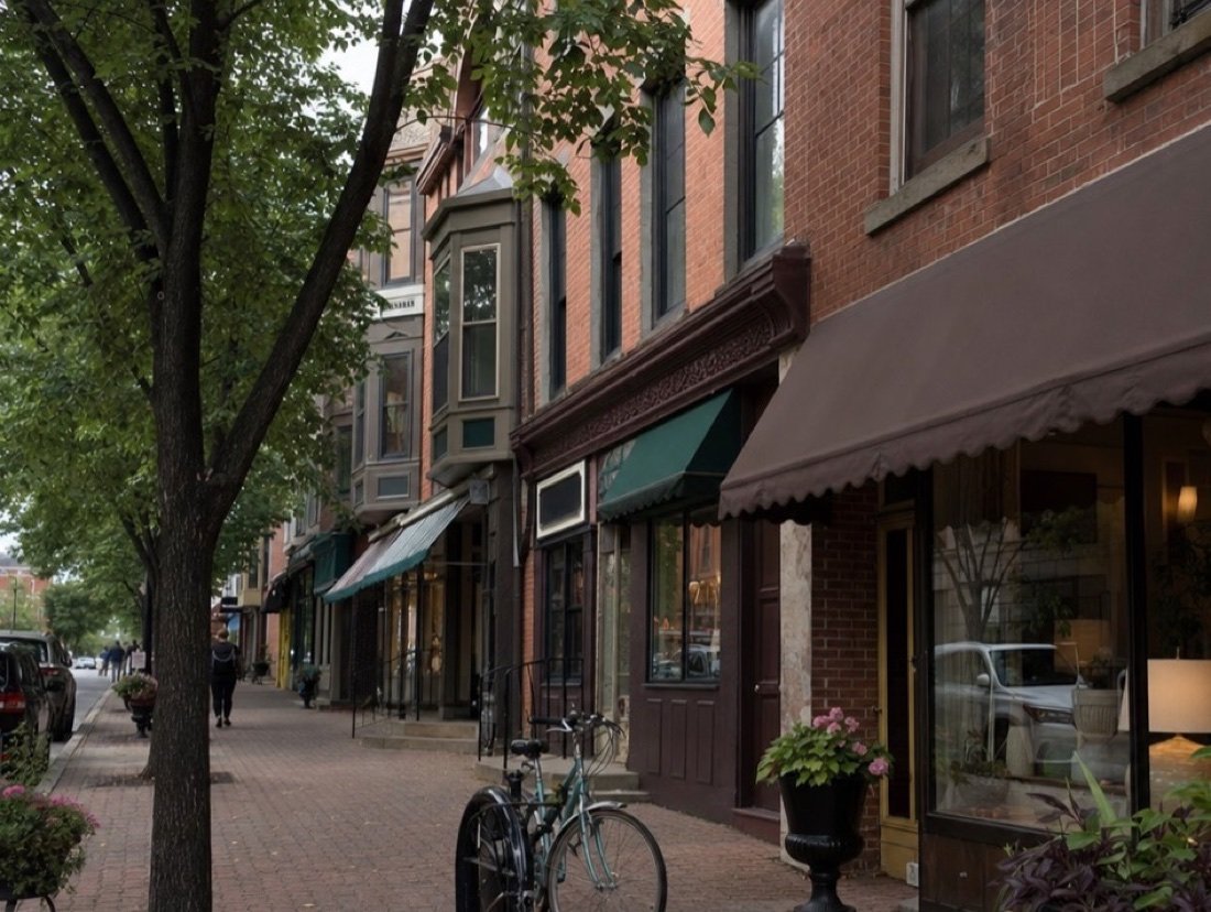 A tree-lined brick sidewalk past three-story red-brick storefronts with bay windows and canvas awnings