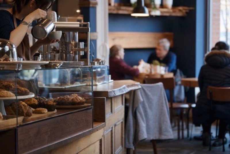 A café counter with baked goods under glass and a barista pouring coffee