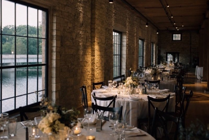 A long stone-walled hall set with round tables and white linen for dinner, tall windows over a river, an events industry setting