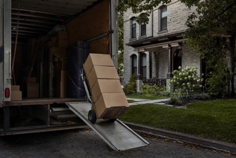 Cardboard cartons piled on a hand truck at the back ramp of a moving van, parked in a driveway