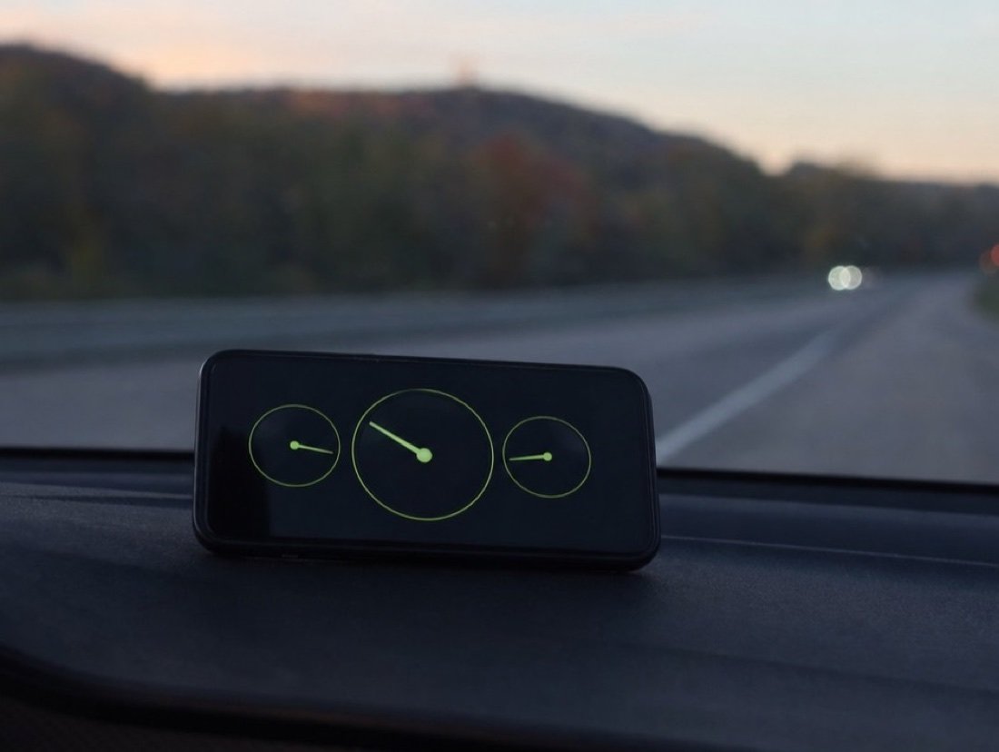 A phone with three green speed gauges mounted on a car dashboard, a highway through wooded hills at dusk