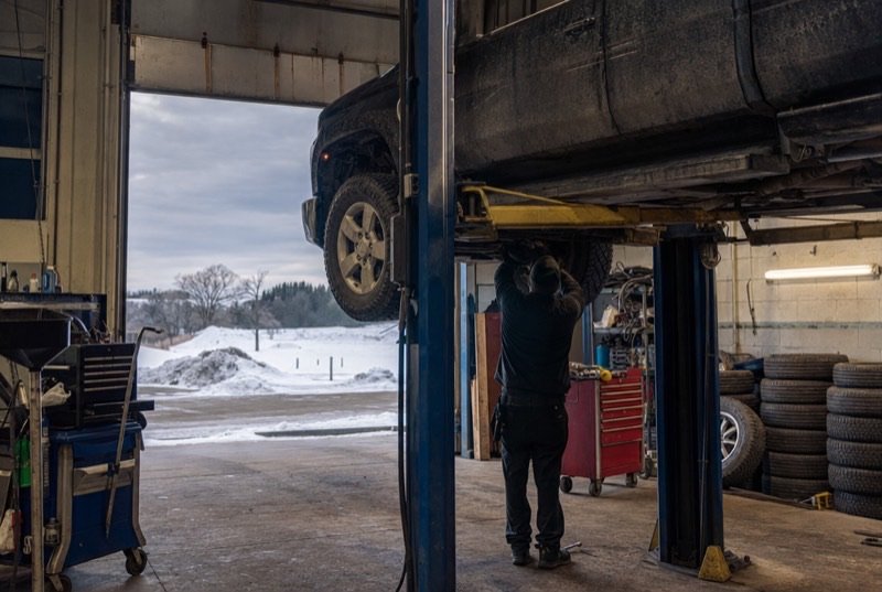 A pickup raised on a garage hoist, winter tires stacked along the wall and snow outside