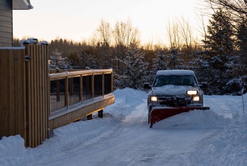 A pickup with a red plow clearing a deep-snow driveway at dusk beside a wooden deck and fence